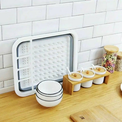 White and gray collapsing dish rack shown folded flat and stored upright on a kitchen counter beside bowls, jars, and spice containers.