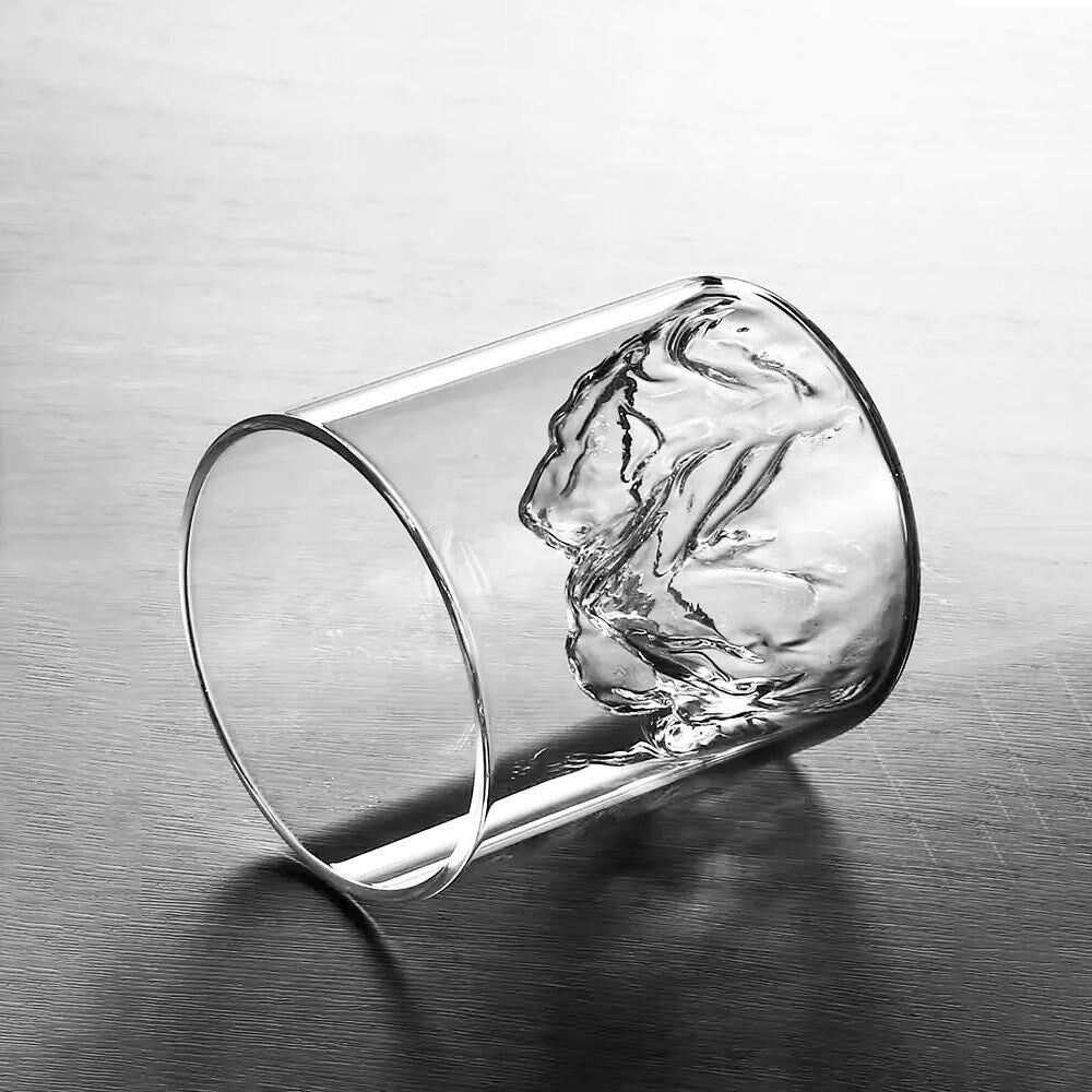 A close-up view of a Colorado mountain whiskey glass lying on its side, showcasing the intricate, sculpted mountain peak design at the base. The clear glass highlights the detailed craftsmanship, reflecting light to emphasize the textured mountain-inspired interior.