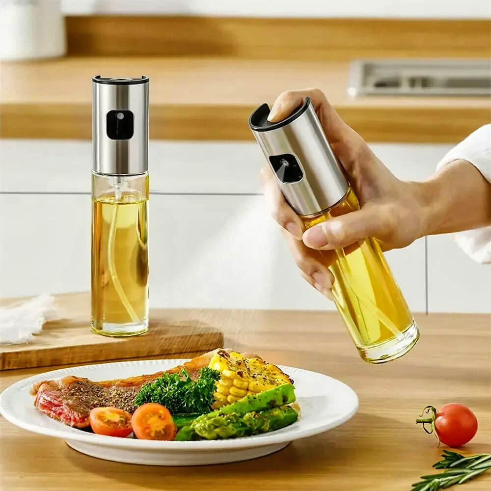 A person is using a cooking oil sprayer bottle to evenly spray oil over a plate of vegetables and grilled meat. Another sprayer bottle stands on the countertop, showcasing its sleek glass body and stainless steel top, perfect for precise and healthy cooking.