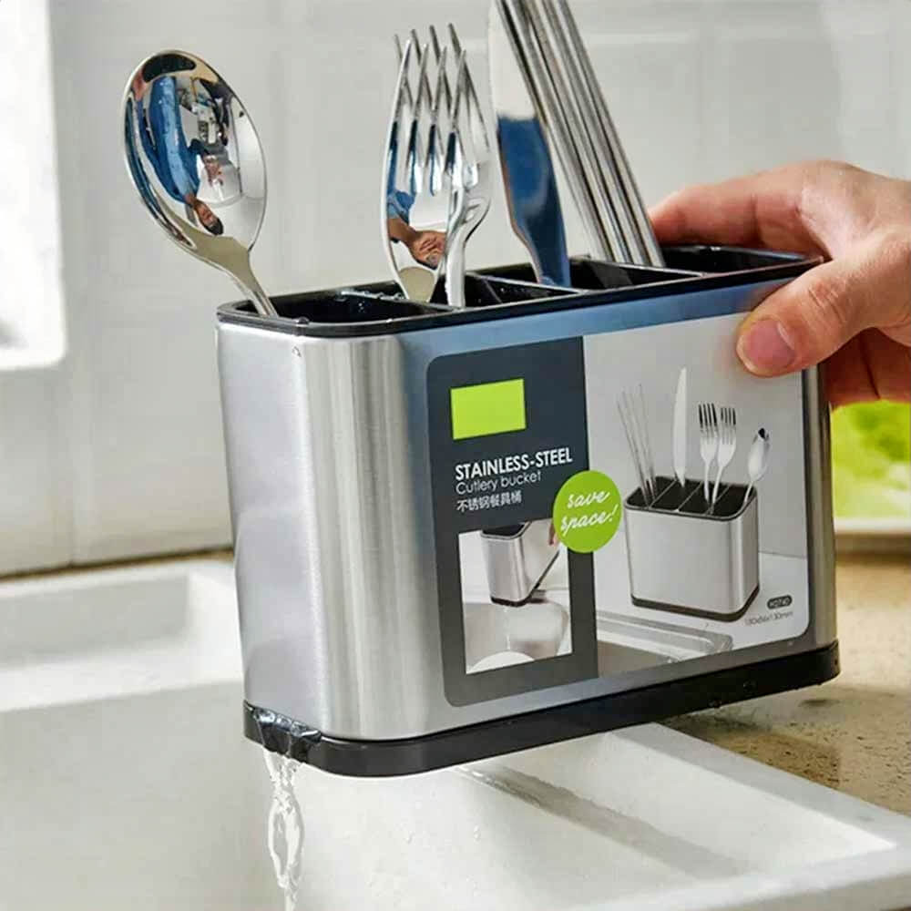Hand holding stainless steel cooking utensil holder with draining base, filled with forks, spoons, and knives over a kitchen sink.