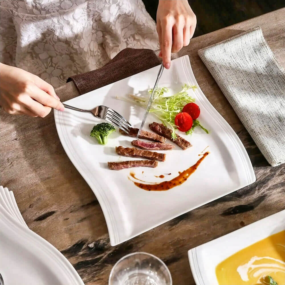 A stylish and contemporary dining setup showcasing Crate and Barrel serving plates. The featured plate has a unique wavy-edge design and is crafted from glossy white ceramic, perfect for elevating meal presentations. It displays an elegant arrangement of steak slices, fresh greens, broccoli, cherry tomatoes, and a drizzle of sauce, highlighting its versatility for various cuisines. A refined choice for sophisticated dining occasions.