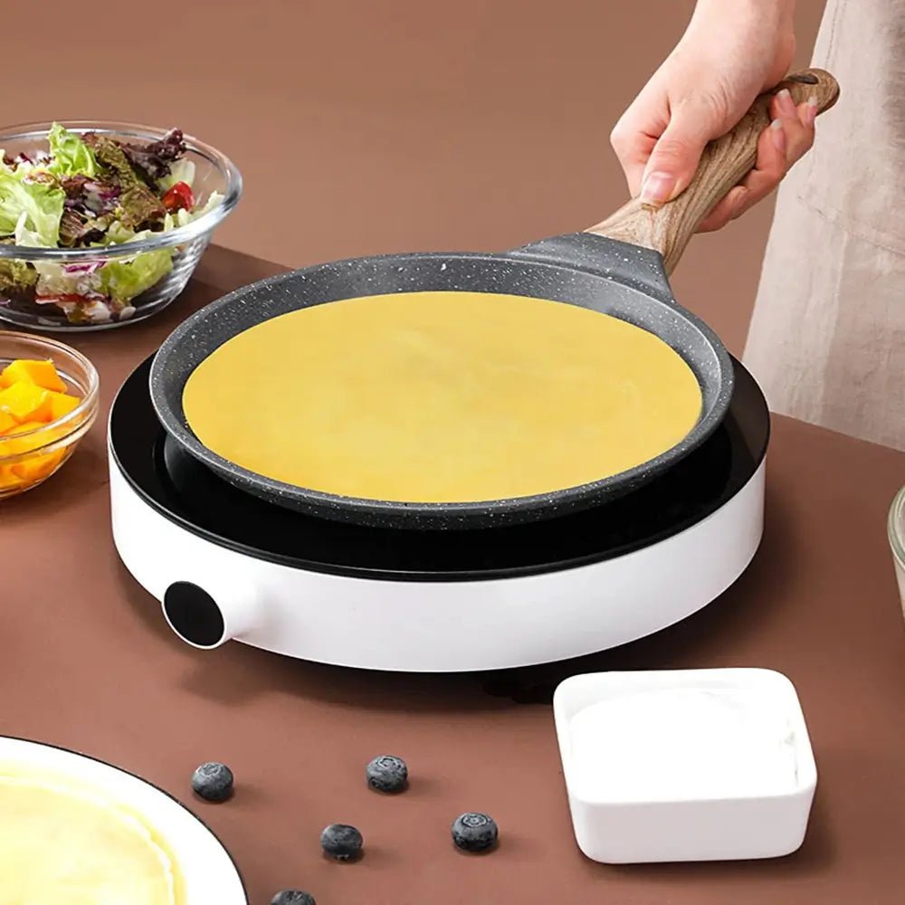 Crepe pan with wooden handle on an induction cooktop, cooking a thin crepe, surrounded by salad, fruit, and yogurt.