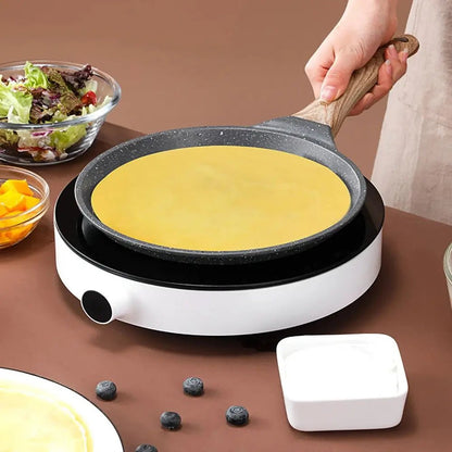 Crepe pan with wooden handle on an induction cooktop, cooking a thin crepe, surrounded by salad, fruit, and yogurt.