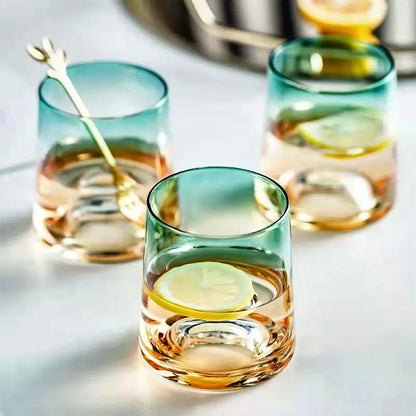 Three gradient green and amber glasses from a crystal carafe and glass set, filled with water and lemon slices, on a white surface.