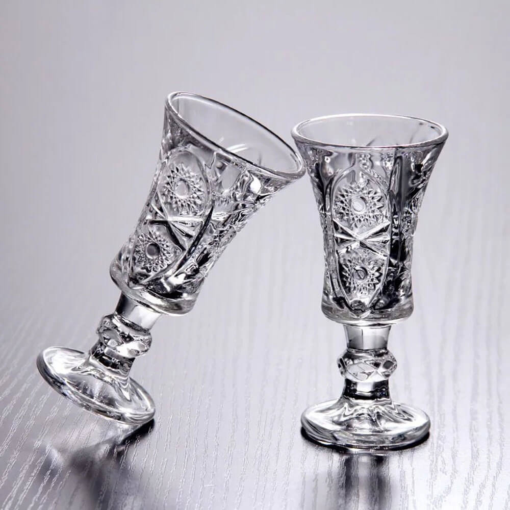 Two intricately designed crystal cocktail glasses are displayed on a reflective surface. The glasses feature detailed floral and geometric patterns, emphasizing their vintage elegance. One glass leans gently against the other, highlighting their delicate craftsmanship and sturdy base.