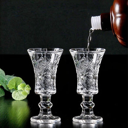 Two crystal glasses with intricate floral and geometric patterns stand elegantly on a reflective black surface. Liquid is being poured into one glass from a brown bottle, emphasizing their use for serving beverages. A touch of greenery in the background adds a fresh, natural contrast to the scene.