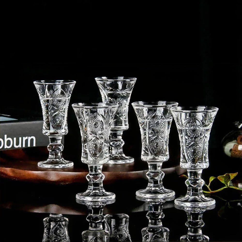 A set of five intricately designed crystal drinking glasses is arranged elegantly on a reflective black surface. The glasses feature ornate floral and geometric patterns, showcasing their vintage charm. A wooden tray and decorative elements in the background enhance the luxurious presentation.