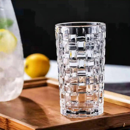 A crystal highball glassware with a geometric lattice design is placed on a wooden tray. The glass is empty, with lemons and a pitcher of ice water visible in the background.