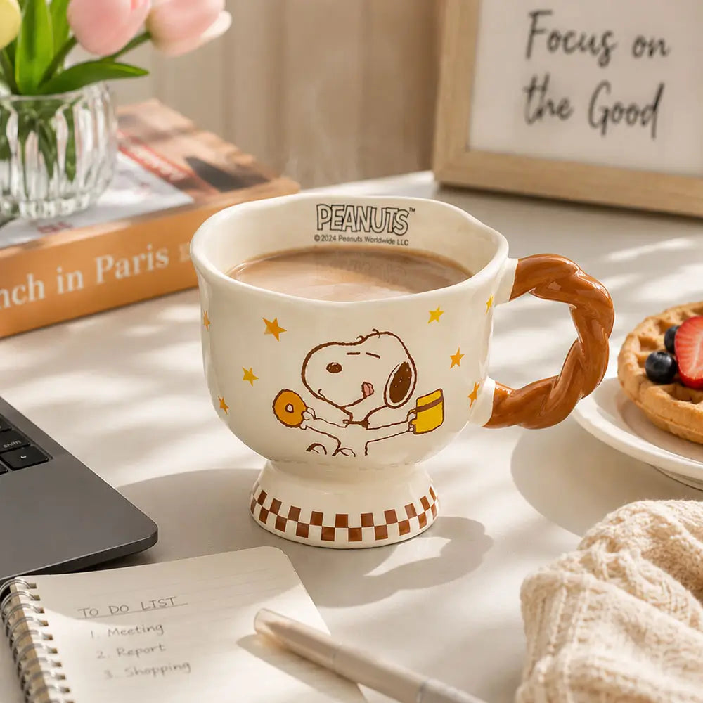 Cartoon coffee mug filled with coffee on a desk beside a laptop, notebook, pen, flowers, and a pastry in a home setting.
