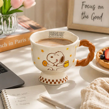 Cartoon coffee mug filled with coffee on a desk beside a laptop, notebook, pen, flowers, and a pastry in a home setting.