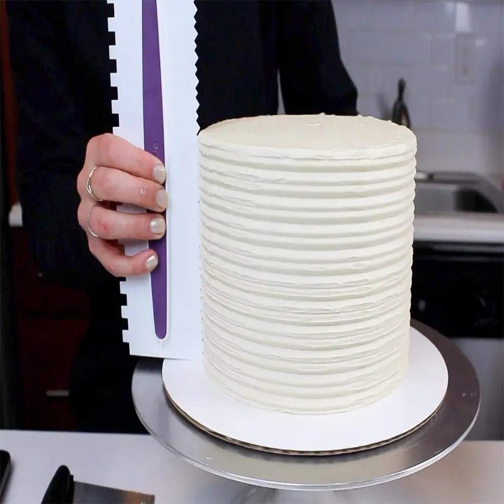 A person uses a decorating comb to create horizontal striped lines on a tall round cake covered in smooth white icing.