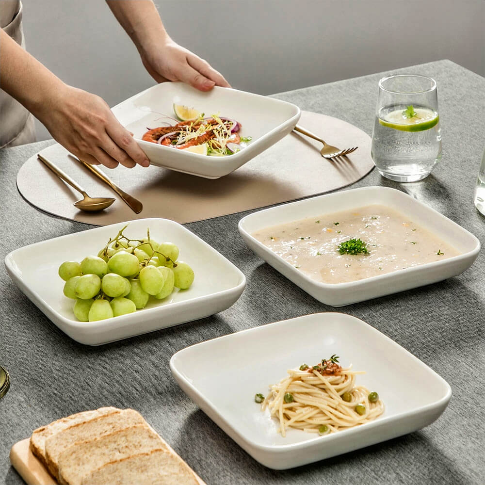 A set of 9.25-inch diameter serving bowls in white porcelain, displayed on a dining table. The bowls contain salad, soup, pasta, and grapes. A person is serving a dish, with gold cutlery, a glass of water with lime, and slices of bread arranged on the table.