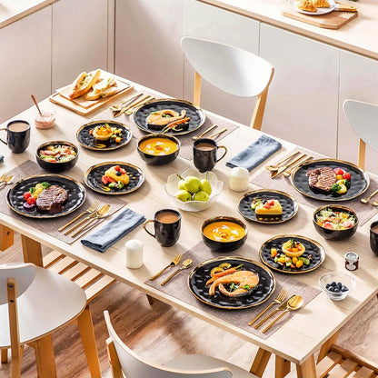 A dining table set with a dinnerware set stoneware featuring glossy black plates, bowls, and mugs accented with gold rims and handles. The table is elegantly arranged with a variety of dishes, including soups, salads, grilled meats, seafood, and desserts, complemented by gold cutlery. Blue napkins, white candles, and a centerpiece with green apples add a stylish and inviting touch to the setting. The natural wood table and minimalist chairs enhance the overall modern and sophisticated atmosphere.