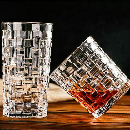 Two double old-fashioned glasses with a geometric lattice pattern are shown. One glass stands upright empty, while the other is tilted and partially filled with an amber liquid, all set on a wooden surface against a dark background.