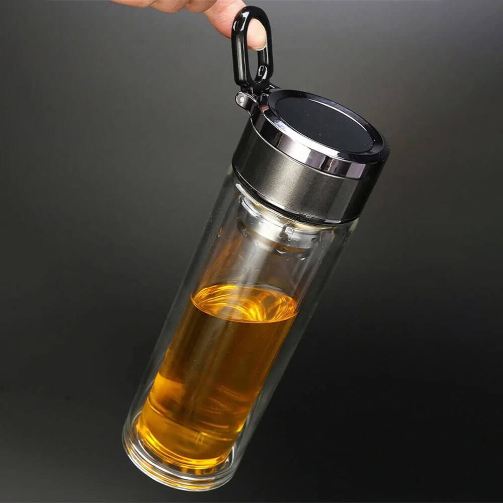 A hand holding a double-wall glass travel tumbler filled with an amber-colored liquid. The tumbler features a sleek, modern design with a metallic lid and a black carabiner clip for easy carrying, set against a dark background.