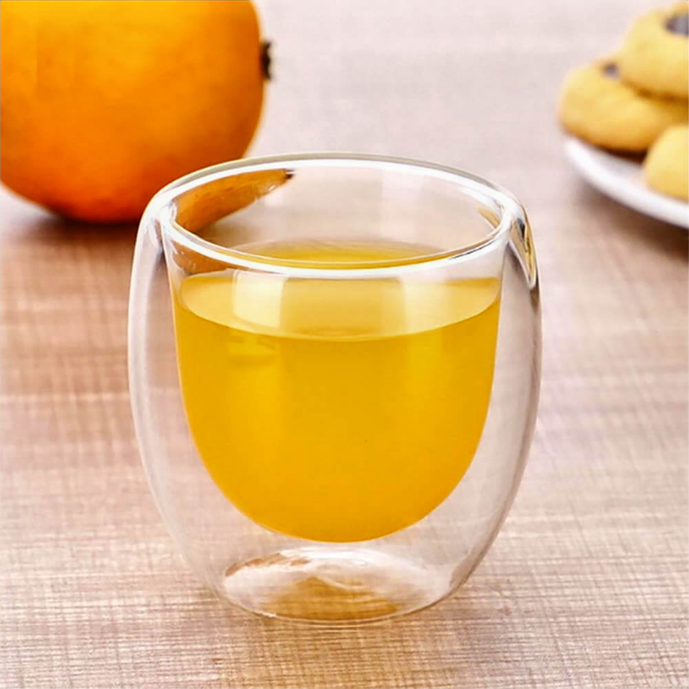 A single double-walled espresso cup made of clear glass is filled with orange juice, placed on a light wooden surface. In the background, there is a whole orange and a plate of cookies, creating a cozy and inviting scene.