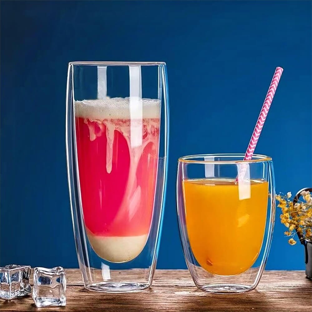 Two double-walled glass coffee tumblers are placed on a wooden surface against a blue background. One tumbler is filled with a pink foamy drink, while the other contains orange juice with a pink straw. Ice cubes and a small vase with flowers are also visible, adding a refreshing and stylish touch.