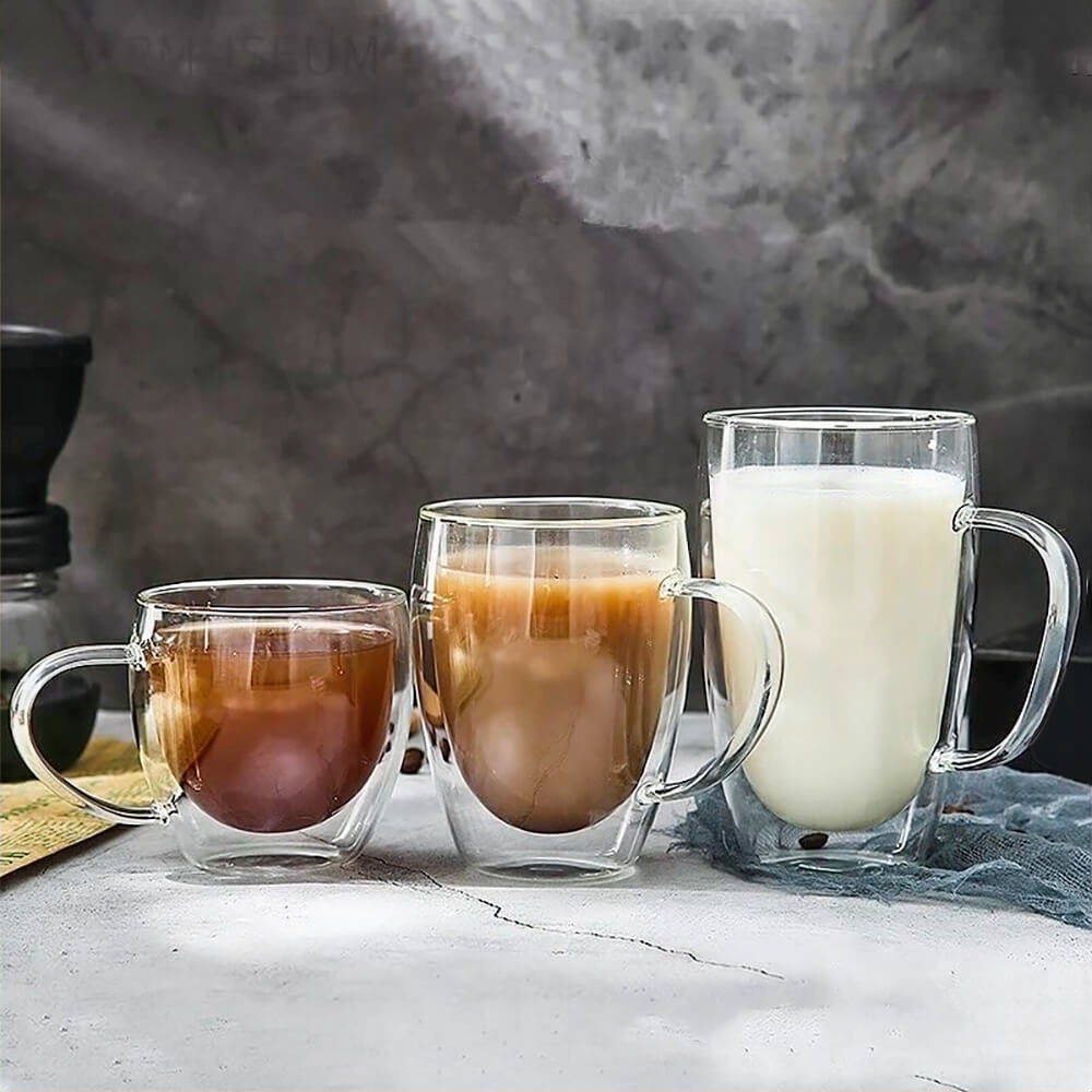 Three double-walled glass tumblers of different sizes, filled with coffee and milk, are placed on a gray surface with a rustic background.