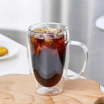 A double-walled glass tumbler filled with iced coffee sits on a wooden surface. The transparent design showcases the beverage and ice cubes, with a clear handle for easy grip.