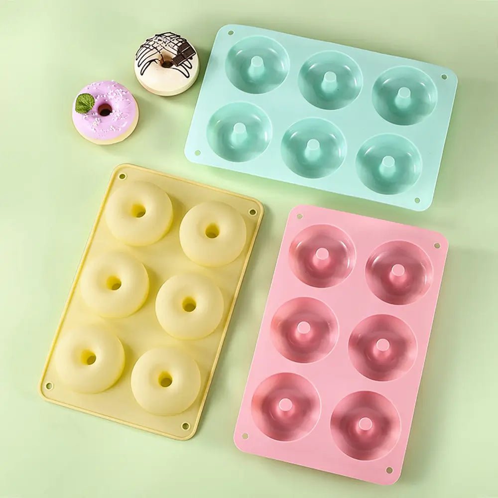 Three silicone doughnut mold pans in pastel pink, yellow, and green, each with six cavities, shown with decorated baked doughnuts nearby.