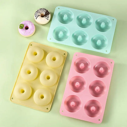 Three silicone doughnut mold pans in pastel pink, yellow, and green, each with six cavities, shown with decorated baked doughnuts nearby.