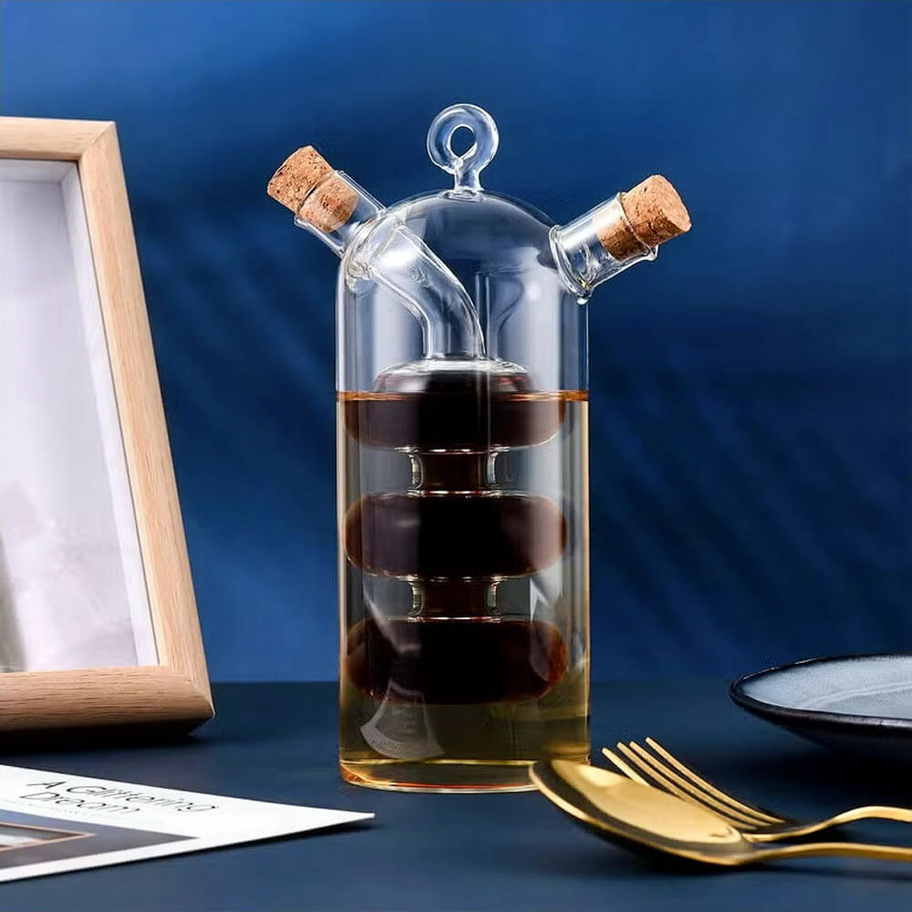 Glass dual oil and vinegar bottle with a stacked design, featuring separate compartments for oil and vinegar. The bottle has two spouts with cork stoppers for controlled pouring and freshness preservation, set against a dark blue background with gold utensils and a framed picture nearby.