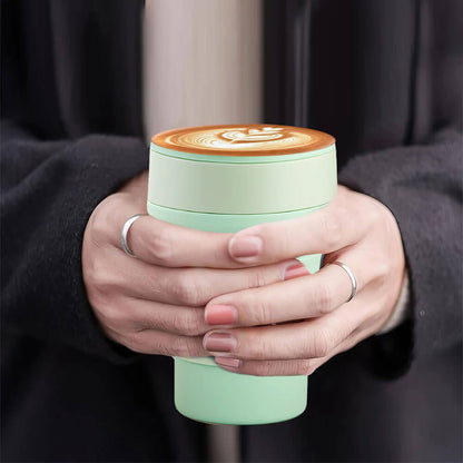 Close-up of hands holding a mint green silicone tumbler filled with coffee, featuring a latte art design on top. The tumbler is compact and stylish, ideal for carrying beverages on the go.