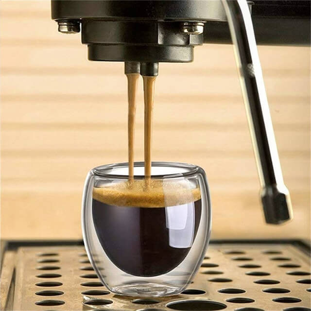 A double-wall espresso cup is placed under an espresso machine, capturing freshly brewed espresso with a rich crema layer. The transparent glass showcases the dark espresso and its creamy top.