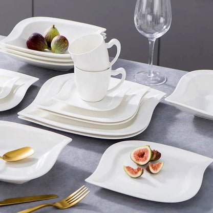 Everyday dinnerware sets with white square and leaf-shaped plates, stacked cups, gold cutlery, figs, and a clear wine glass on a table.