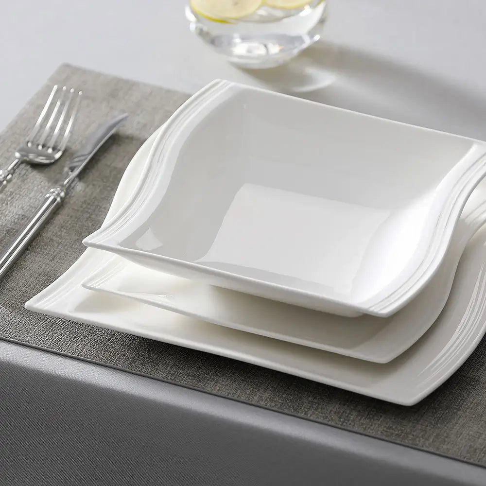 White everyday porcelain dinnerware plates and bowl with undulating rims stacked on a gray placemat next to silverware.
