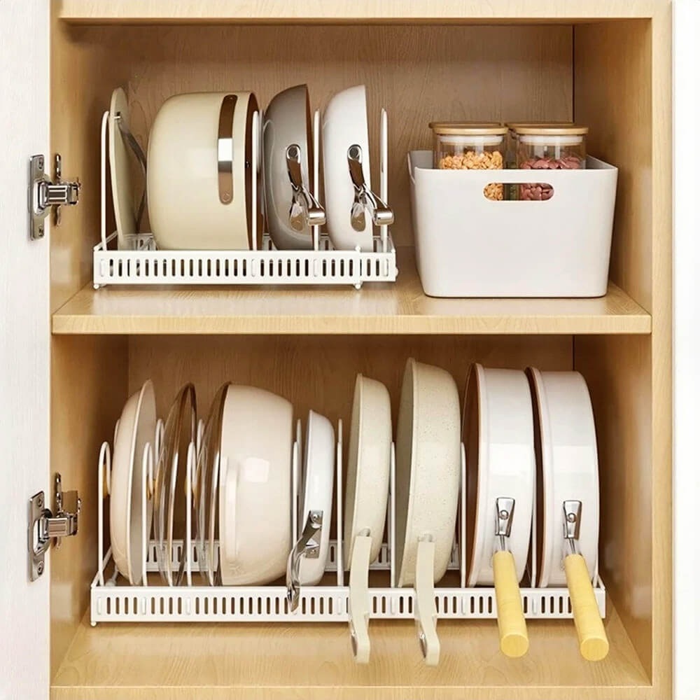 
A neatly organized kitchen cabinet with two shelves features an expandable pan rack on each level. The racks securely hold pots, pans, and lids in an upright position for easy access. The upper shelf also includes a white storage bin containing jars of food items.