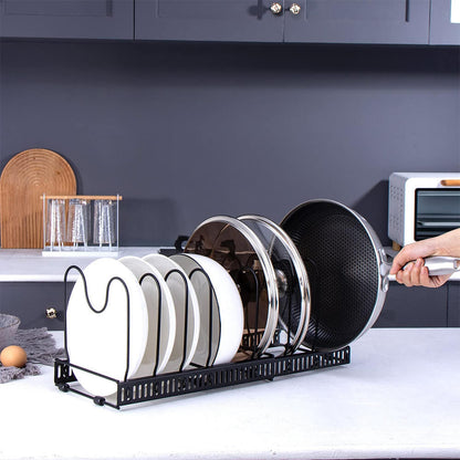 A black expandable pot and pan organizer rack is placed on a white countertop. It holds white plates, glass lids, and a frying pan upright. Dark gray cabinets and a wooden cutting board are visible in the background. A person is removing the frying pan.