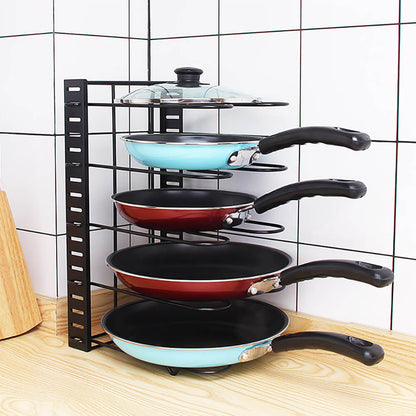 A black expandable pot rack is placed on a wooden countertop. It holds four frying pans, two blue and two red, stacked horizontally, along with a glass lid on the top shelf. The setup is against a white tiled wall.