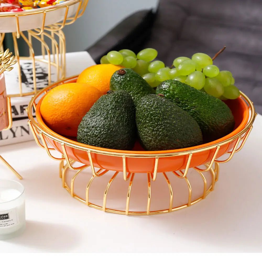 Extra-large pedestal fruit bowl holding avocados, oranges, and grapes in a ceramic dish with gold wire stand on table.