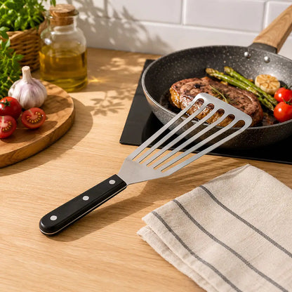 Fish turner spatula displayed on a kitchen countertop beside a frying pan with cooked food and cooking ingredients.