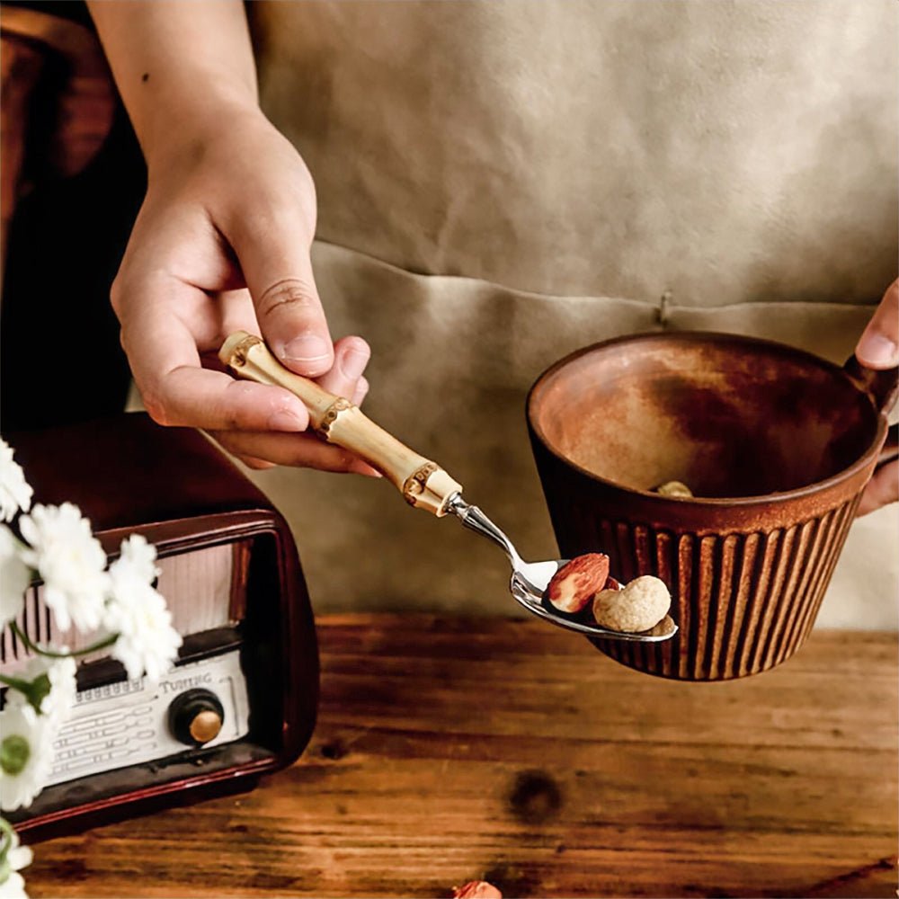 A hand holds a bamboo-handled spoon with dried fruits over a rustic ceramic mug on a wooden table.