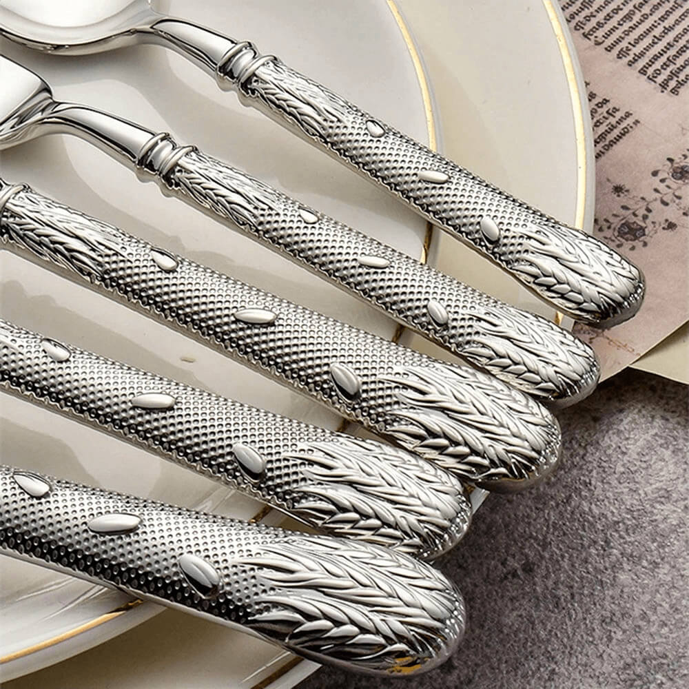 Close-up view of flatware collections arranged on a plate, showcasing detailed handles with embossed leaf and floral patterns. The polished stainless steel finish enhances the intricate design, highlighting the fine craftsmanship and luxurious appeal of the flatware.