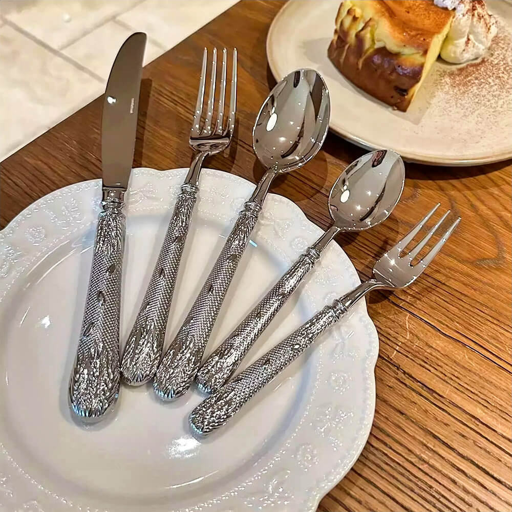 A floral flatware set displayed on a white decorative plate, featuring a knife, fork, tablespoon, teaspoon, and salad fork. Each piece is crafted from polished stainless steel with intricately embossed floral and leaf patterns on the handles. The flatware is elegantly arranged, highlighting its detailed design, with a dessert plate and cake slice in the background.