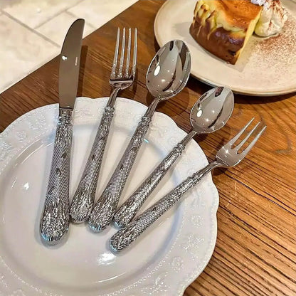 Set of textured silver cutlery with floral handles placed on a white plate, dessert slice visible in the background on wooden table.