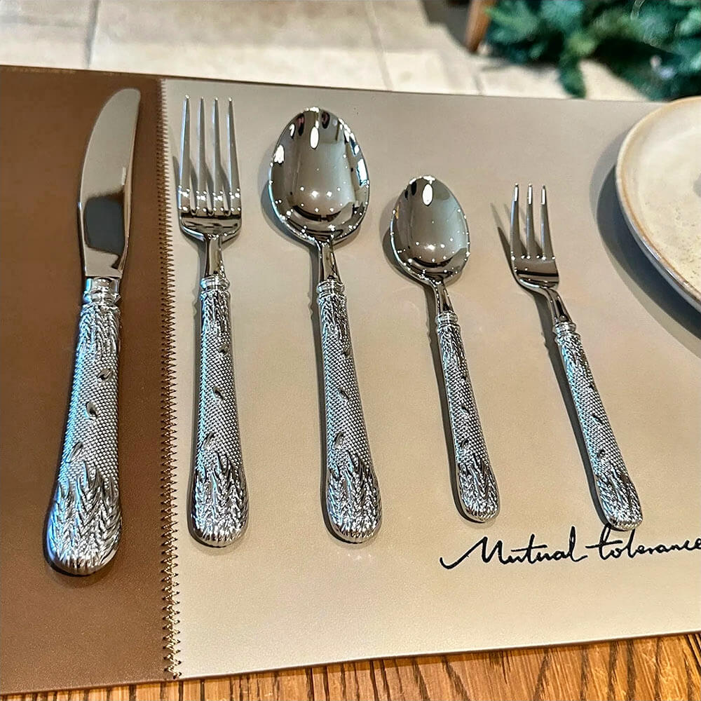 A floral flatware set neatly arranged on a beige placemat, featuring a knife, fork, tablespoon, teaspoon, and salad fork. Each piece showcases polished stainless steel with intricately embossed floral and leaf patterns on the handles, blending elegance and functionality. The setting is minimal and sophisticated, highlighting the luxurious design of the flatware.