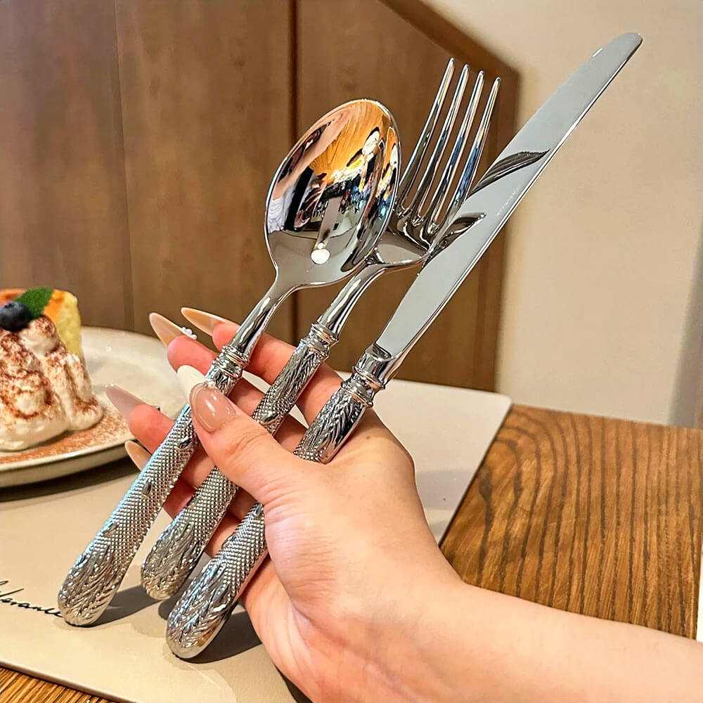 A hand holding a set of floral flatware, including a knife, fork, and spoon. The polished stainless steel utensils feature intricately embossed handles with floral and leaf patterns, reflecting a sophisticated design. A dessert plate with a slice of cake is visible in the background, adding to the elegant dining setting.