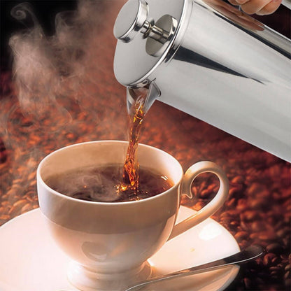 French press stainless steel pouring hot coffee into a white cup with saucer, surrounded by coffee beans.
