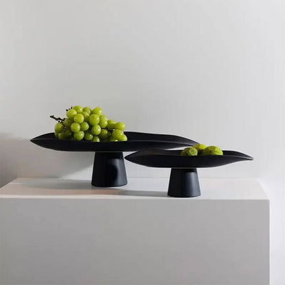 Two elegant black ceramic fruit bowls with a minimalist design, each featuring a wide, curved surface and a pedestal base, beautifully displaying green fruits.