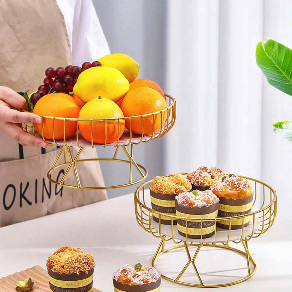 Two elegant pedestal bowls with gold wire frames displayed on a table. The larger bowl holds fresh fruits, including oranges, lemons, and grapes, while the smaller bowl contains delicious muffins. A person is seen lifting the fruit bowl, adding functionality and charm to the scene.