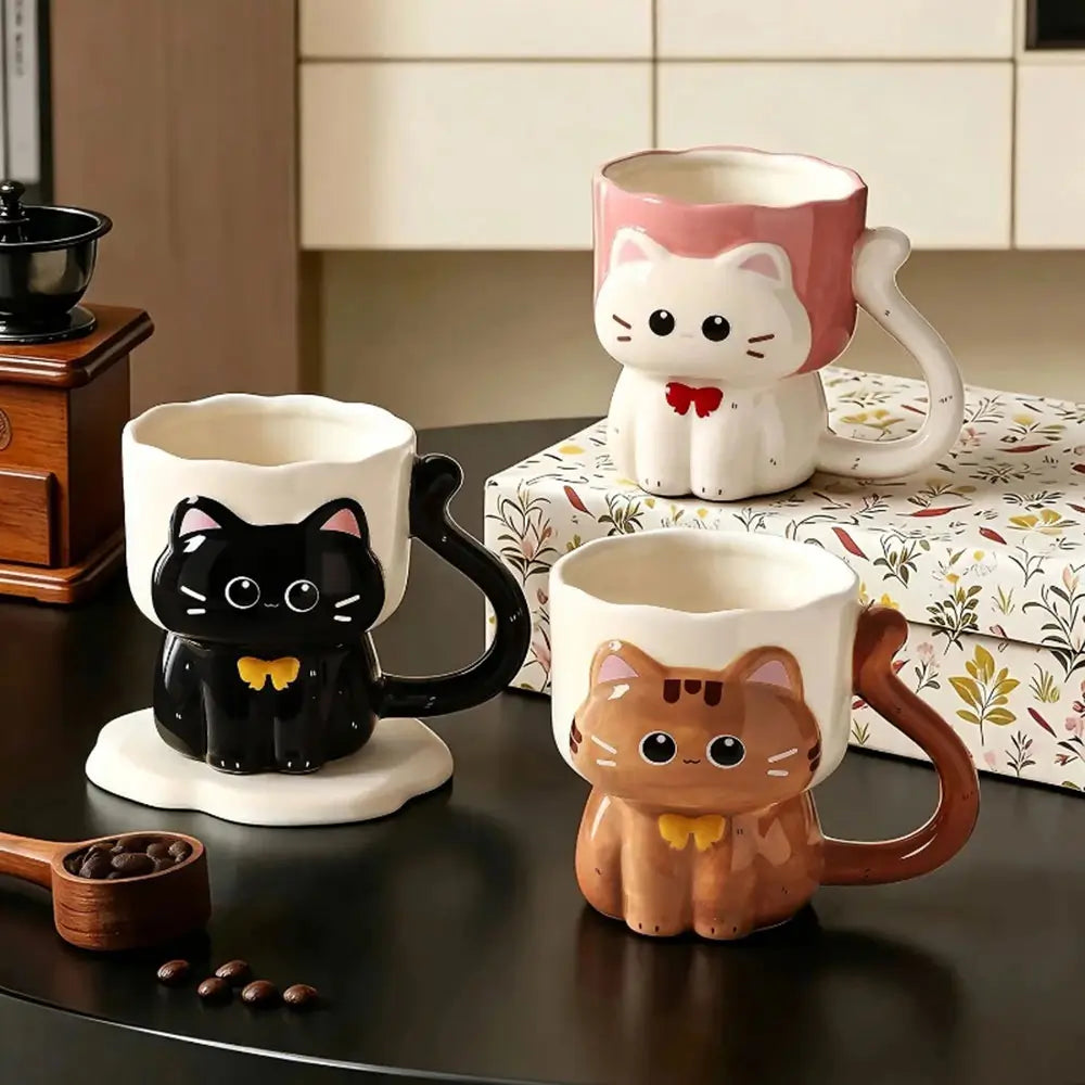 Three ceramic cat-shaped mugs with handles are arranged on a dark tabletop in a kitchen setting. One mug is black with a yellow bow tie and white base, one is white with a pink top and white base, and one is brown with a yellow bow tie and white base.