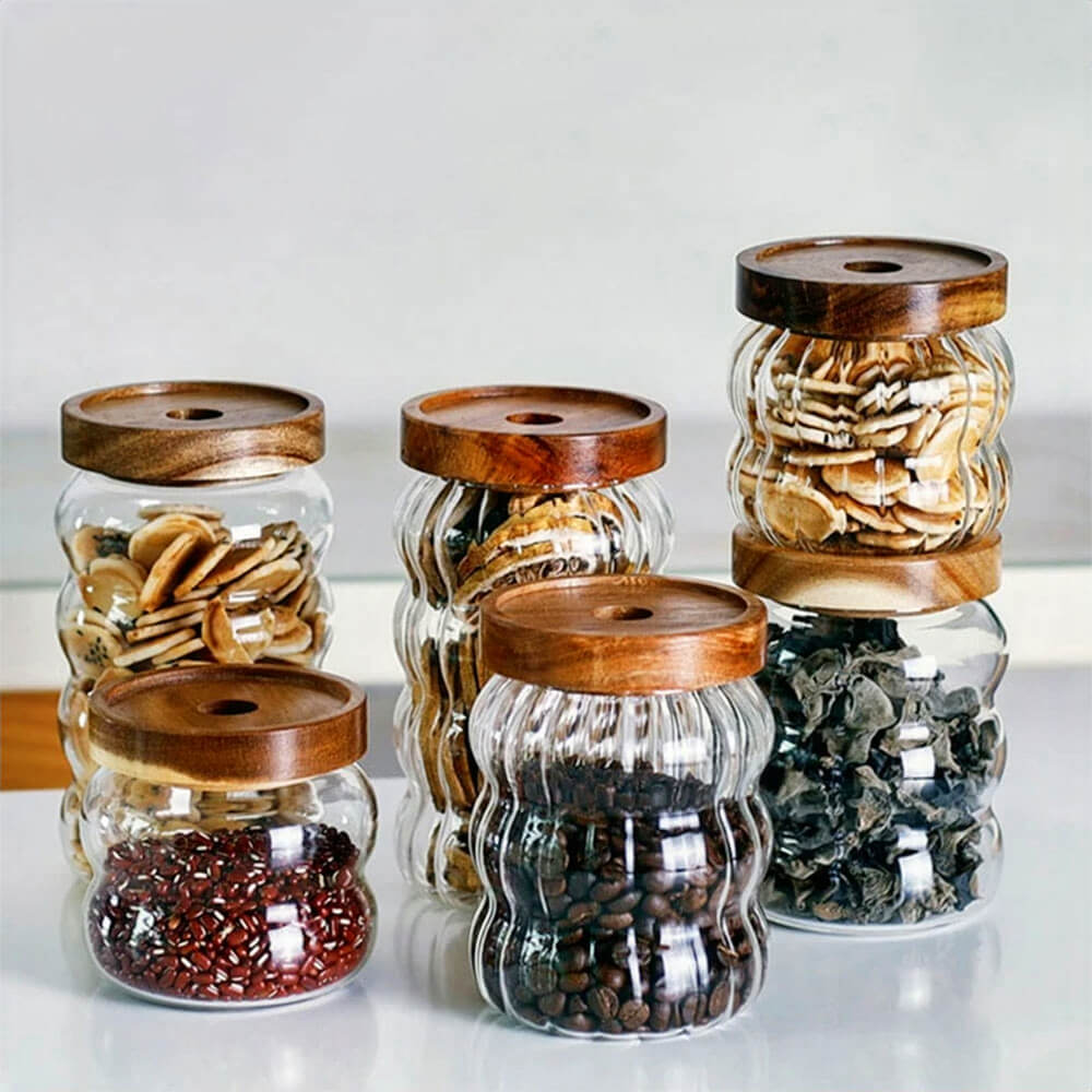 A set of six glass canisters in varying sizes is arranged on a countertop. Each jar features clear ripple-textured glass and a round dark wooden lid with a small central hole. The canisters are filled with assorted dried foods, including coffee beans, red beans, mushrooms, and cookies.