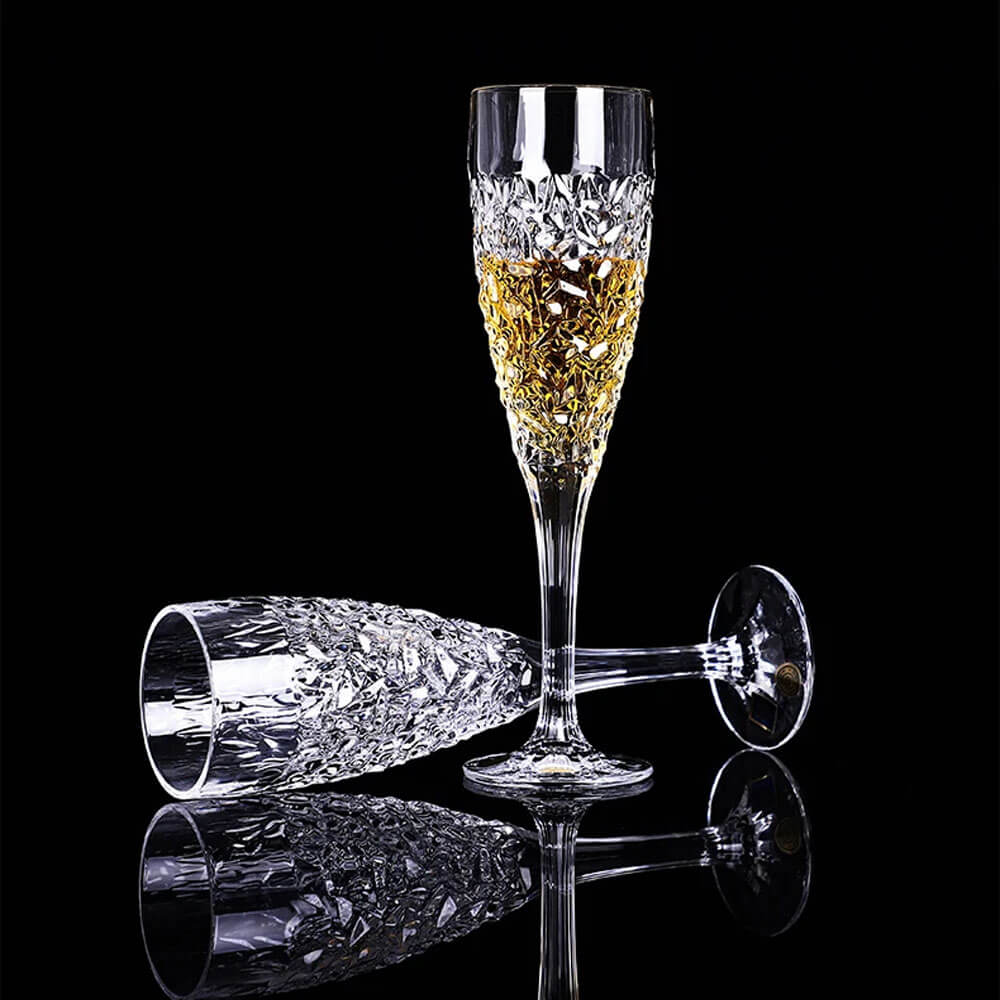 Two intricately designed glass flute glasses, one standing upright filled with a golden liquid and the other lying down, both featuring faceted patterns, displayed against a black background.