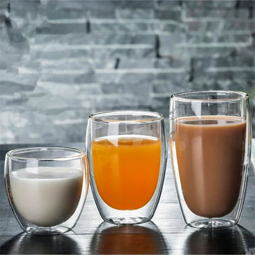 Three glass coffee tumblers of varying sizes are displayed, each filled with a different beverage—milk, orange juice, and coffee. The tumblers feature a double-walled design and are placed on a dark surface with a gray textured background, highlighting their modern and versatile style.