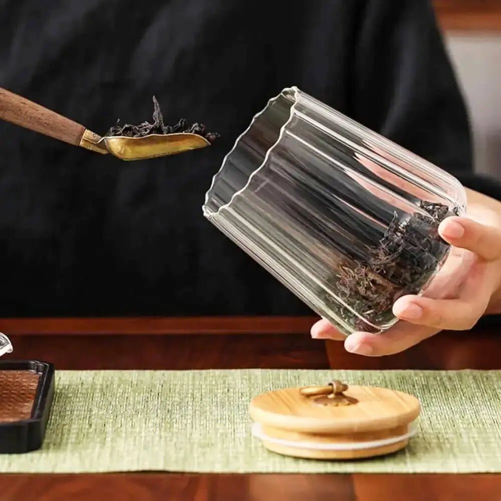 Person holding a glass container with tea leaves, using a wooden scoop, on a tea-making setup.