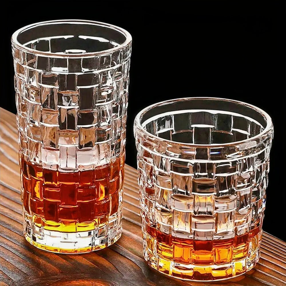 Two old-fashioned glasses with a geometric lattice design are shown on a wooden surface. Both are partially filled with an amber liquid, contrasting beautifully with the clear, textured glass. The setting features a dark background, emphasizing the intricate pattern and reflective quality of the glasses.